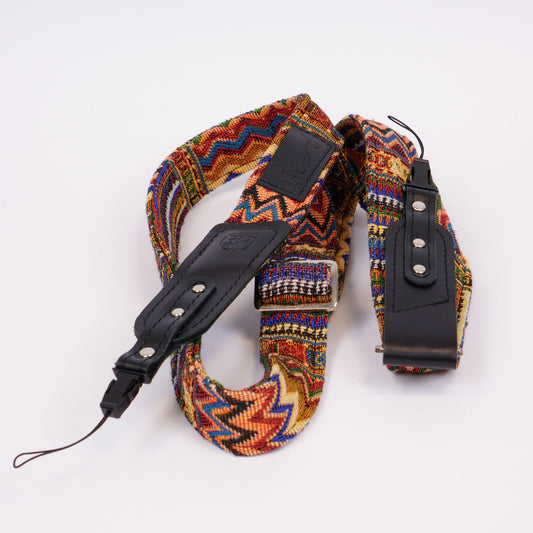 A multicolor patterned camera strap with black leather accents and silver metal hardware, photographed on a clean white background.
