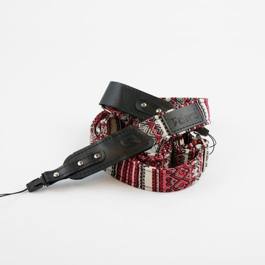 A coiled camera strap featuring a red, black, and white patterned woven fabric body with black leather accents and silver metal rivets, on a plain white background.