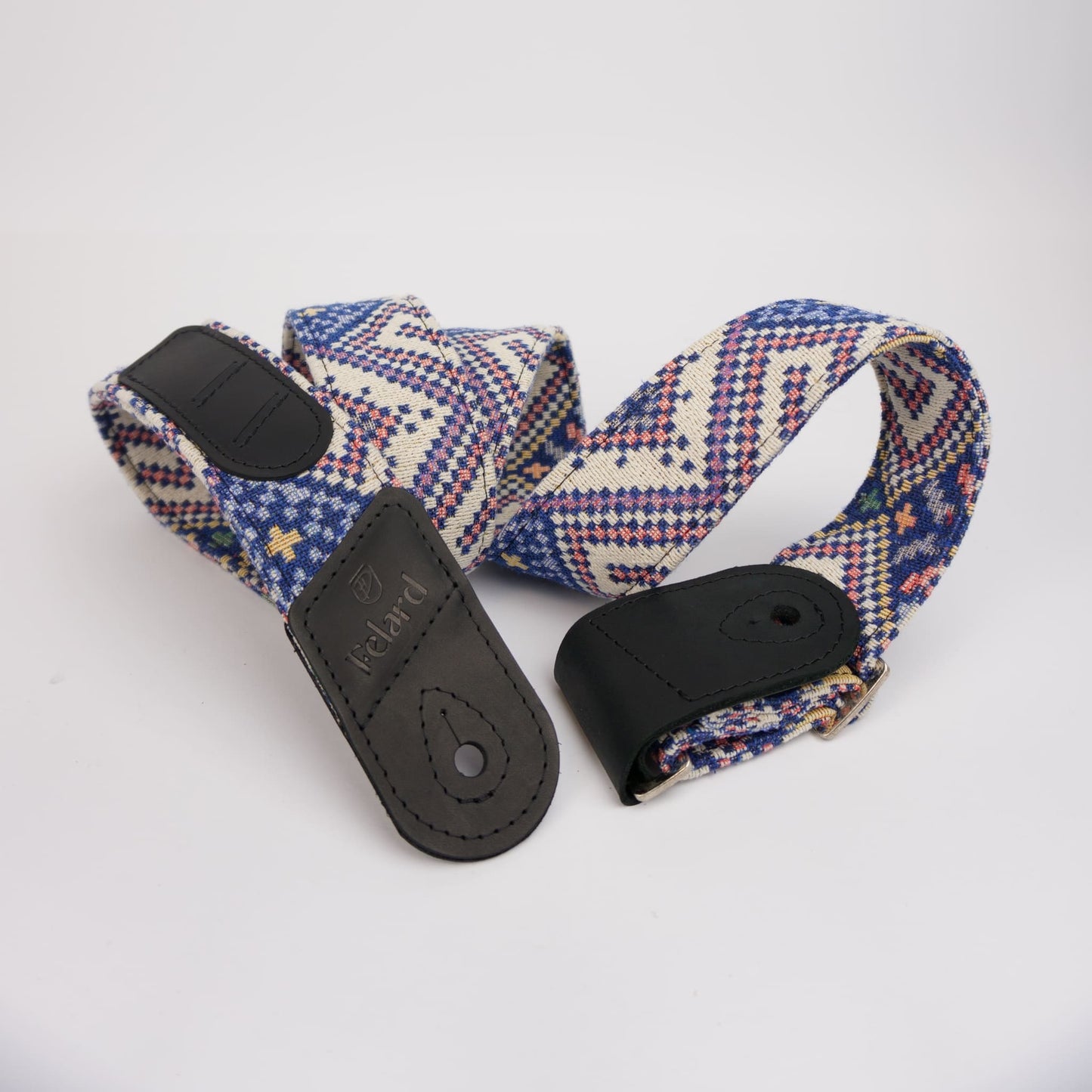 A guitar strap with a blue, red, and cream geometric patterned fabric body and black leather ends, one of which is embossed with the brand name 'Felard'.