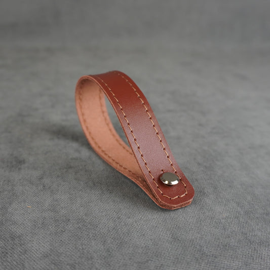 A cognac smooth leather strap headstock adapter with a single silver metal rivet, photographed on a textured gray background.