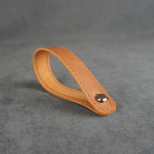 A light tan leather strap headstock adapter with a single silver metal rivet, photographed on a textured gray background.