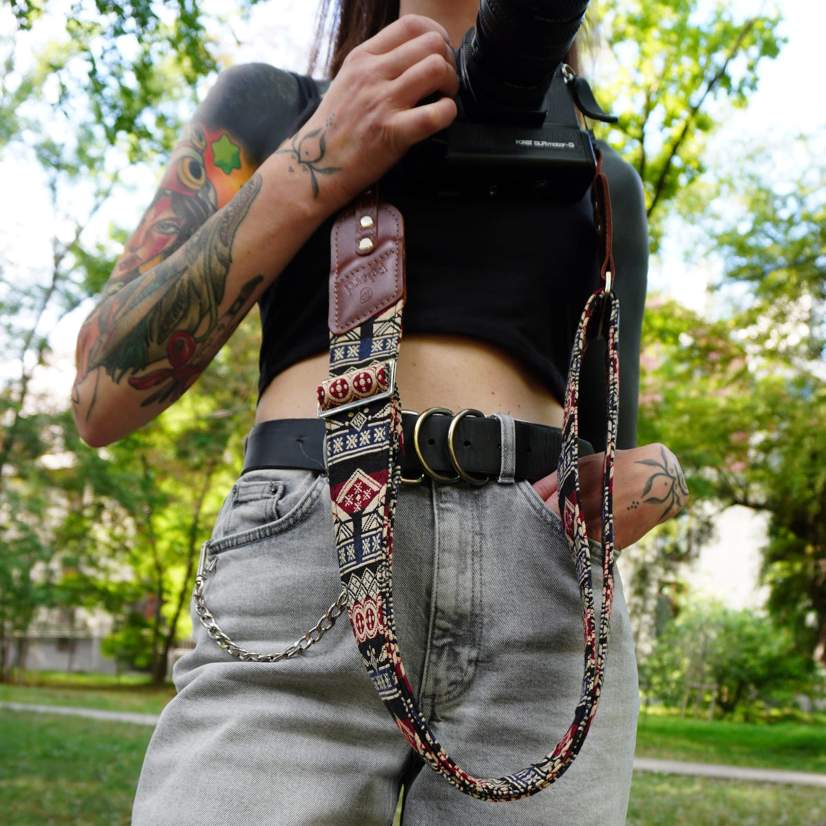 A person outdoors wearing a patterned camera strap with brown leather trim over a black top and light gray jeans, holding a camera.