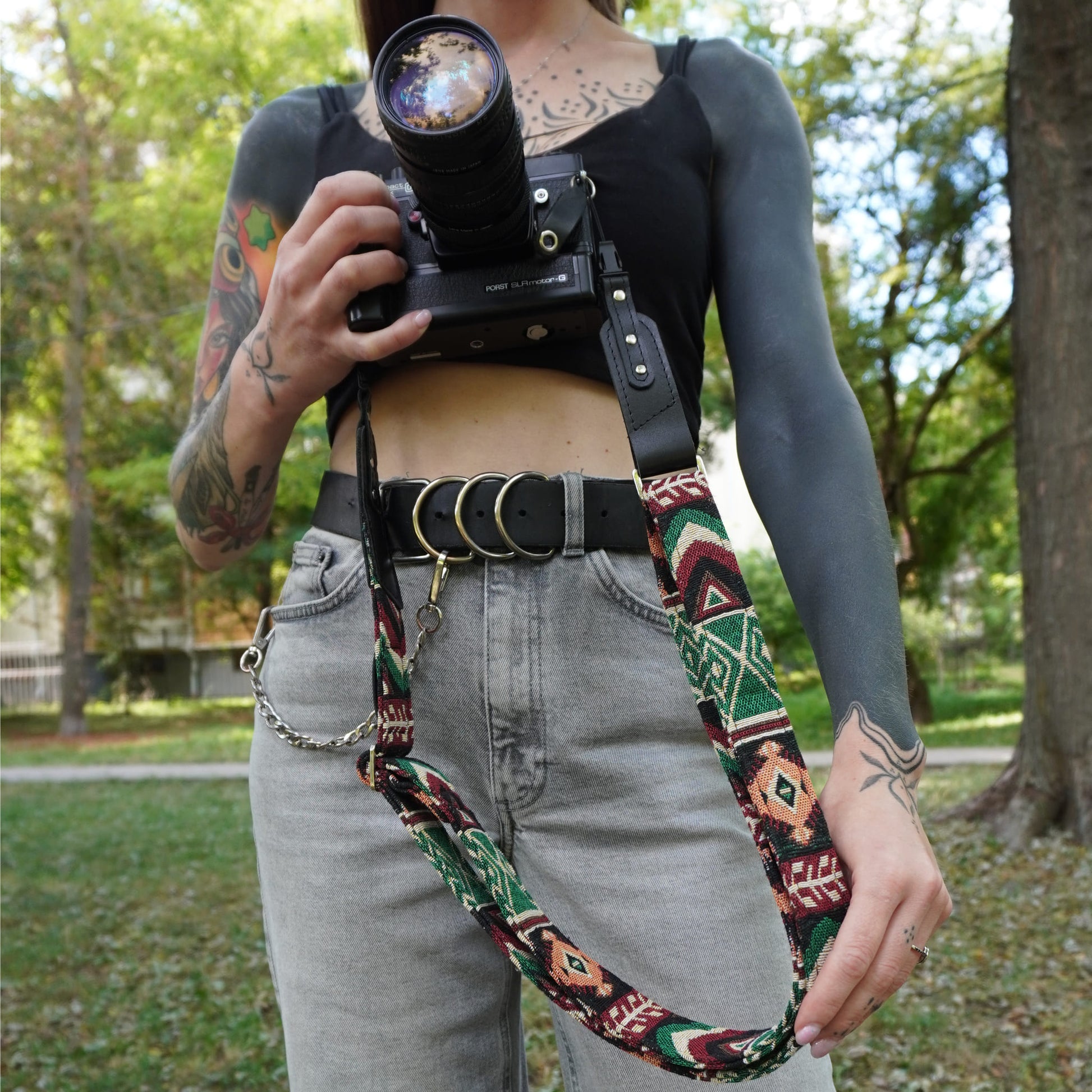 A close-up lifestyle shot of a woman wearing a bohemian-style woven camera strap with black leather accents. The strap features a multicolor geometric pattern in green, red, and beige, with black leather leather sections and silver-toned metal hardware.
