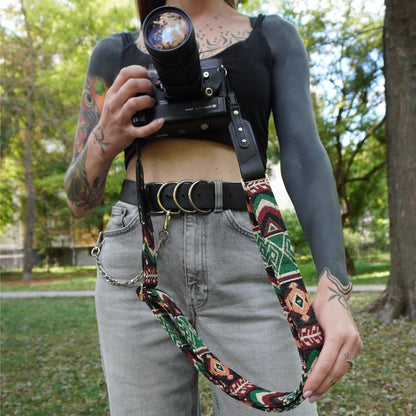A close-up lifestyle shot of a woman wearing a bohemian-style woven camera strap with black leather accents. The strap features a multicolor geometric pattern in green, red, and beige, with black leather leather sections and silver-toned metal hardware.