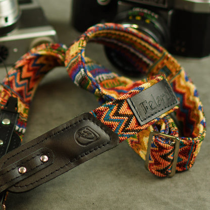 A close-up of a colorful woven camera strap with black leather and metal rivets, shown on a gray surface with vintage cameras blurred in the background.