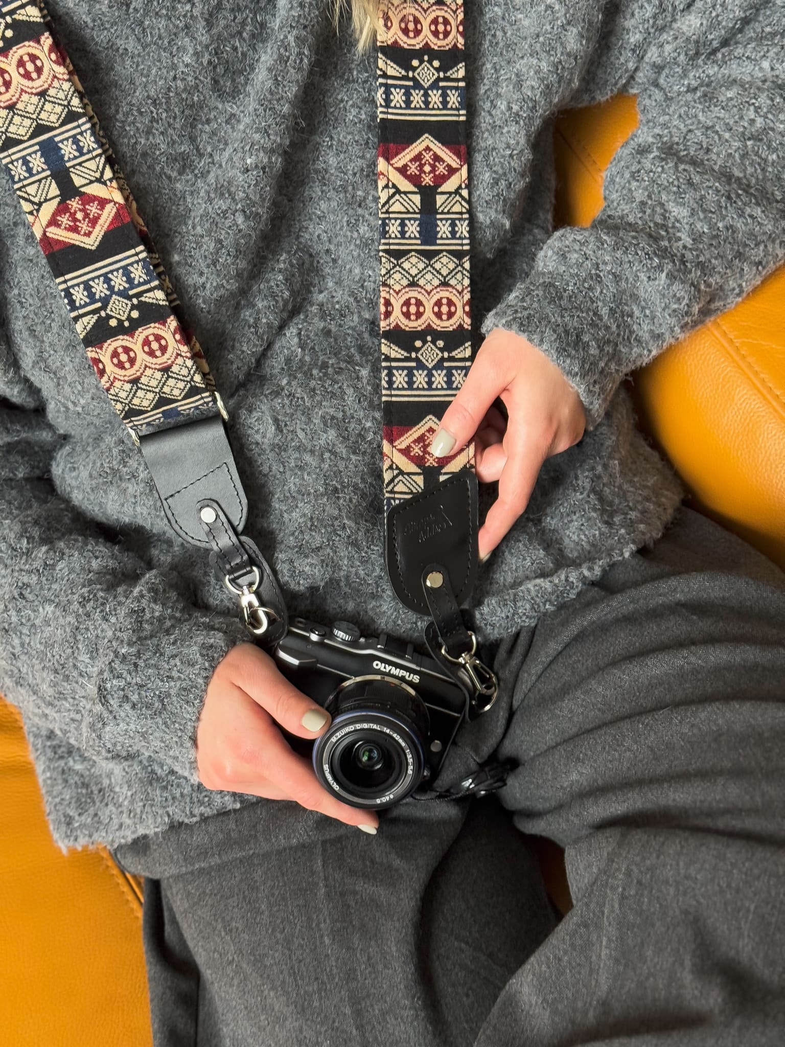 A close-up lifestyle shot of a person holding a black camera with a patterned camera strap. The strap features a Southwestern geometric design in red, beige, and black, with a black leather loop at the end.