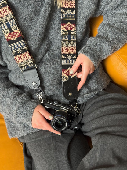 A close-up lifestyle shot of a person holding a black camera with a patterned camera strap. The strap features a Southwestern geometric design in red, beige, and black, with a black leather loop at the end.
