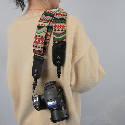 A person wearing a cream knit sweater carries a camera with a patterned woven strap. The strap features a Southwestern geometric design in red, green, beige, and black, with black leather sections.