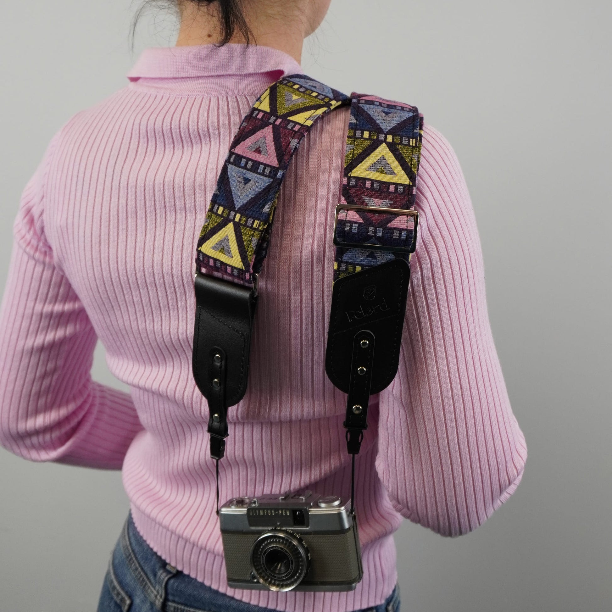 A person viewed from behind with a patterned camera strap draped over the shoulders and holding a silver camera. The strap has a multicolor geometric print with black leather ends.