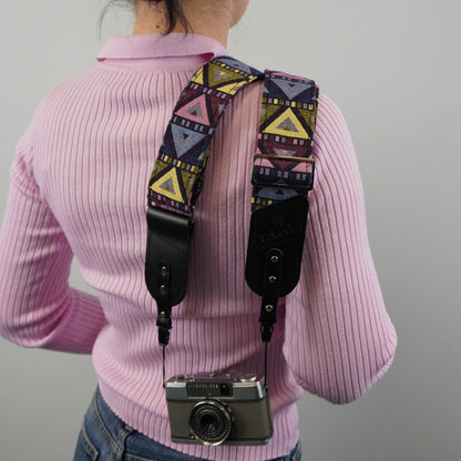 A person viewed from behind with a patterned camera strap draped over the shoulders and holding a silver camera. The strap has a multicolor geometric print with black leather ends.