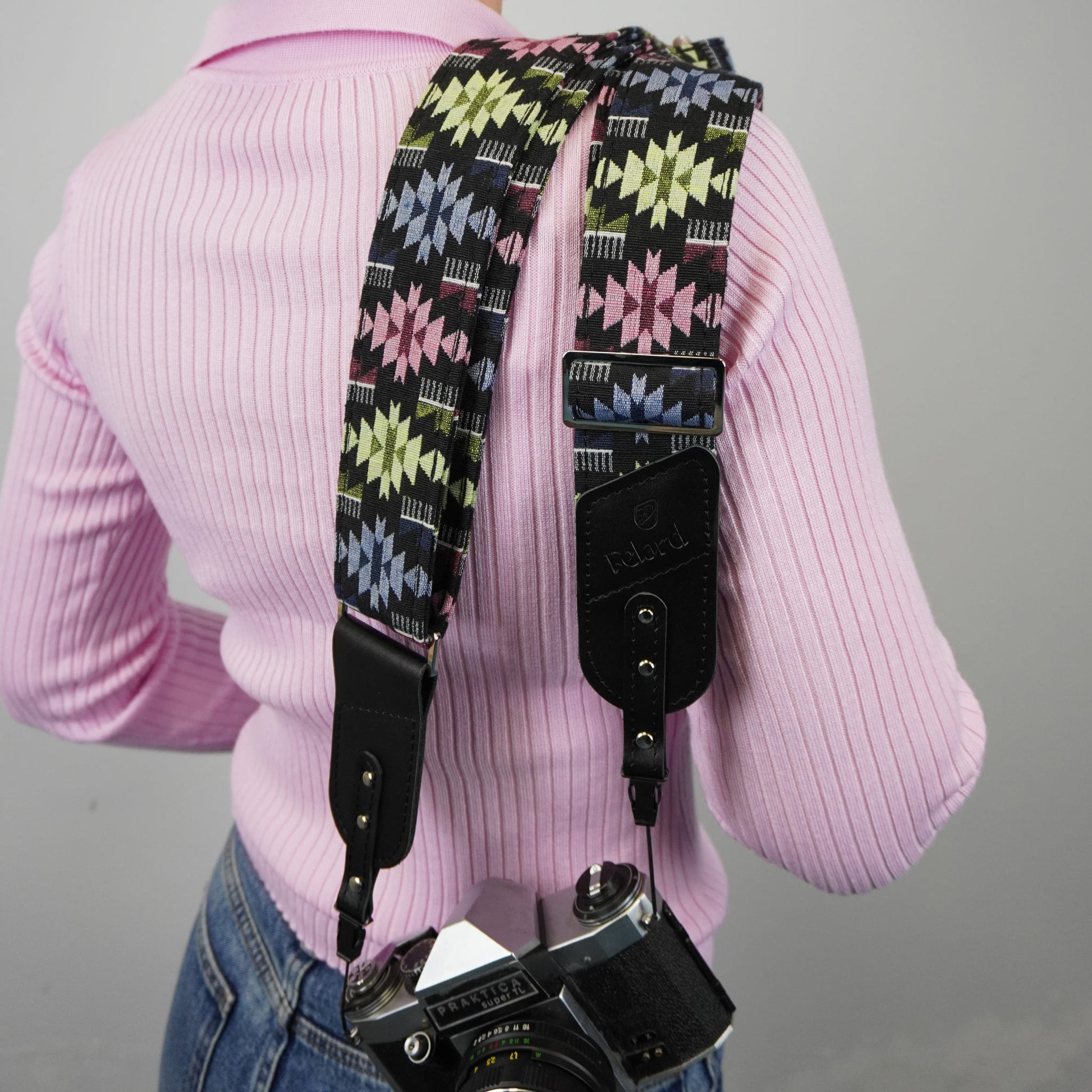 A person shown from the back with a patterned camera strap hanging over the shoulders and a camera at the waist. The strap has a black base with geometric starburst motifs in pink, yellow, blue, and white, plus black leather hardware.