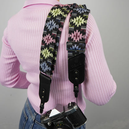 A person shown from the back with a patterned camera strap hanging over the shoulders and a camera at the waist. The strap has a black base with geometric starburst motifs in pink, yellow, blue, and white, plus black leather hardware.