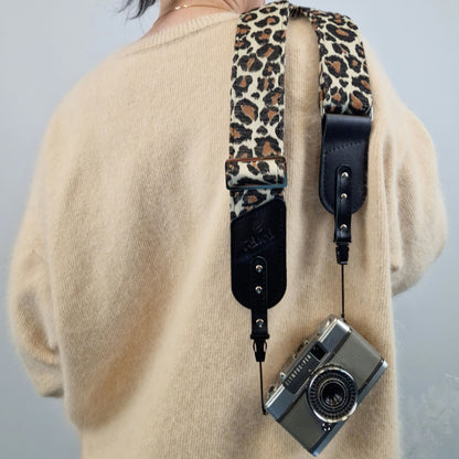 A person is shown from the back, with a leopard-print camera strap draped over their shoulders and holding a silver compact camera. The strap features black leather-like end pieces and side hardware.