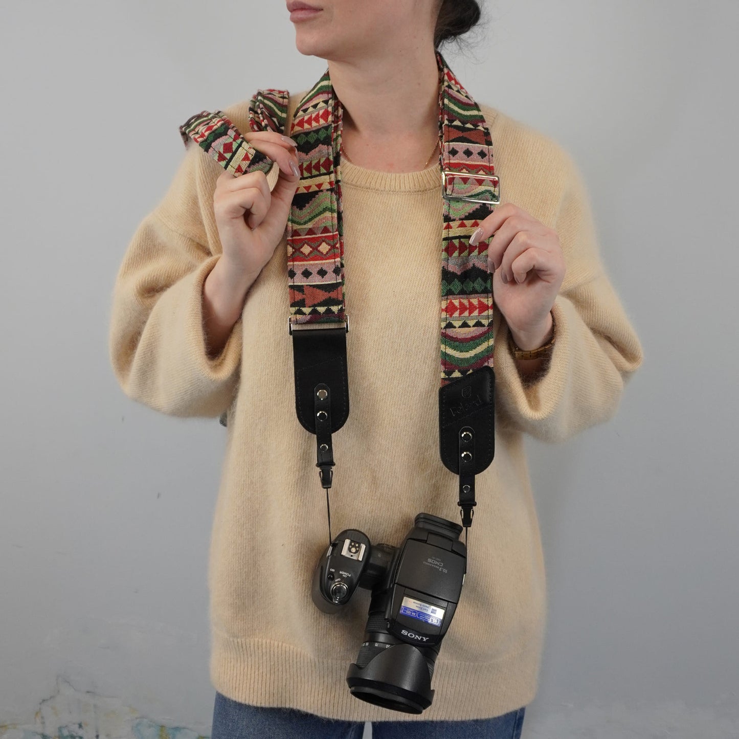 A person wearing a beige sweater models a colorful patterned camera strap with black leather accents, holding a Sony camera with the strap attached against a plain light gray background.