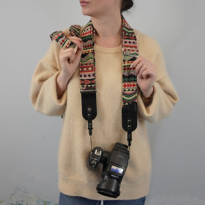 A person wearing a beige sweater models a colorful patterned camera strap with black leather accents, holding a Sony camera with the strap attached against a plain light gray background.