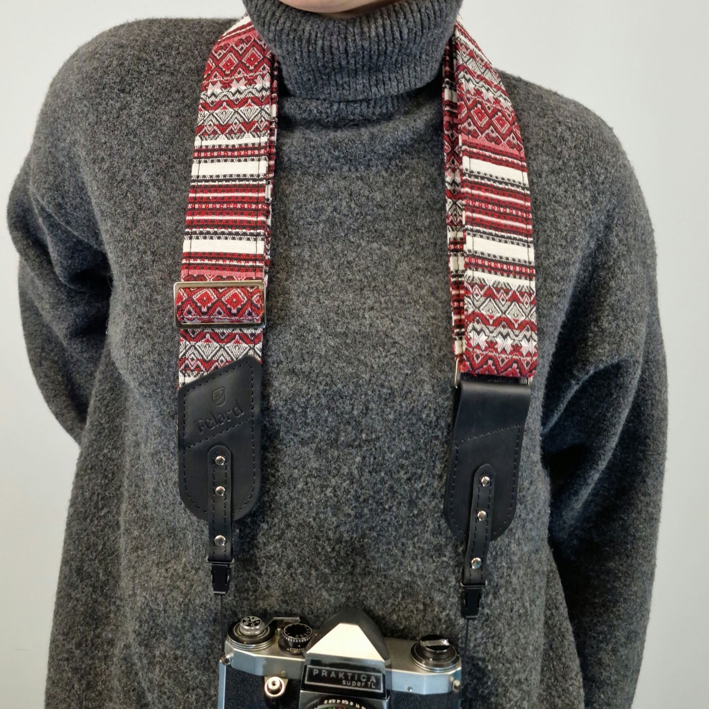 A close-up of a person wearing a patterned camera strap with black leather ends. A vintage camera is partially visible hanging from the strap against a plain light background.