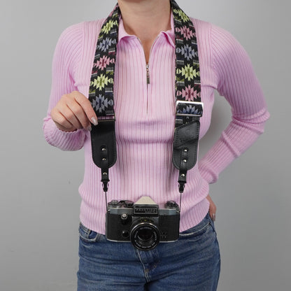A person models a camera strap with a black and multicolor geometric pattern and black leather ends, holding a vintage black camera against a plain gray background.