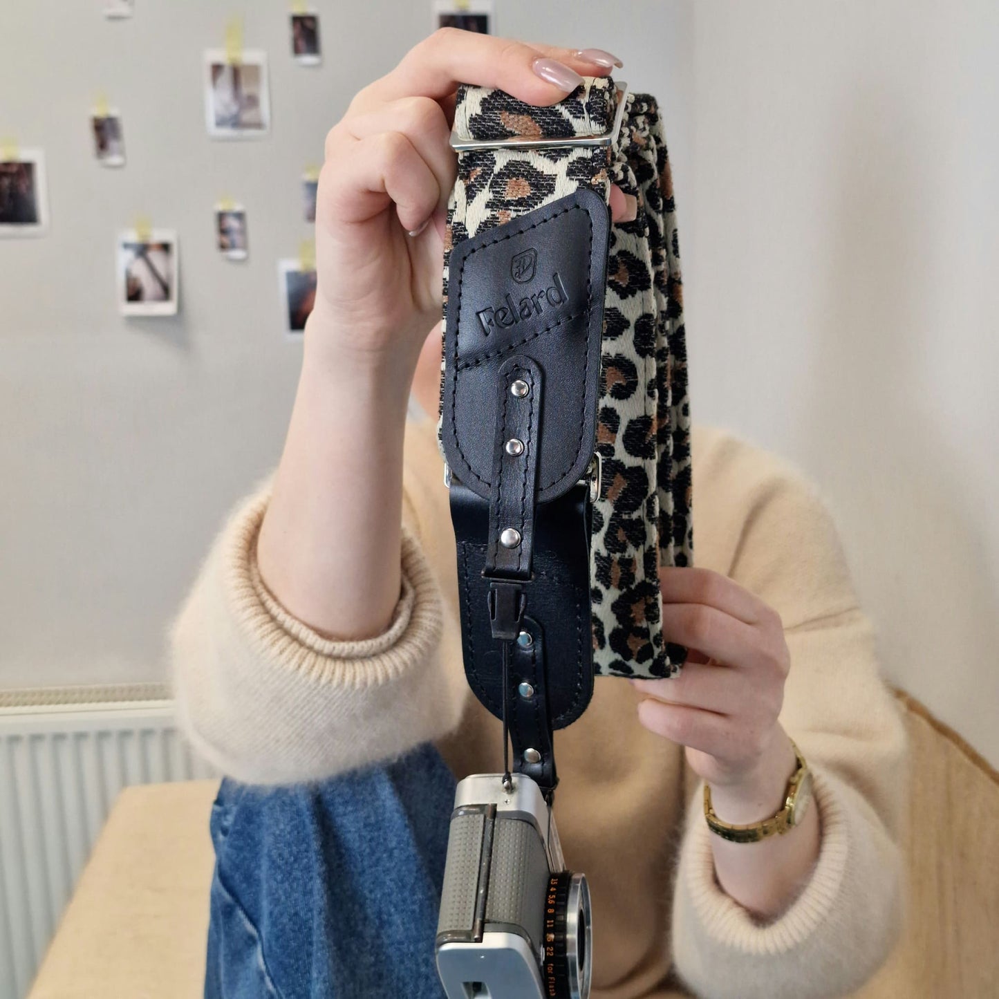 A person holds up a camera strap with a leopard print pattern and a black leather accent, with a vintage camera hanging from it.