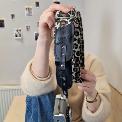 A person holds up a camera strap with a leopard print pattern and a black leather accent, with a vintage camera hanging from it.