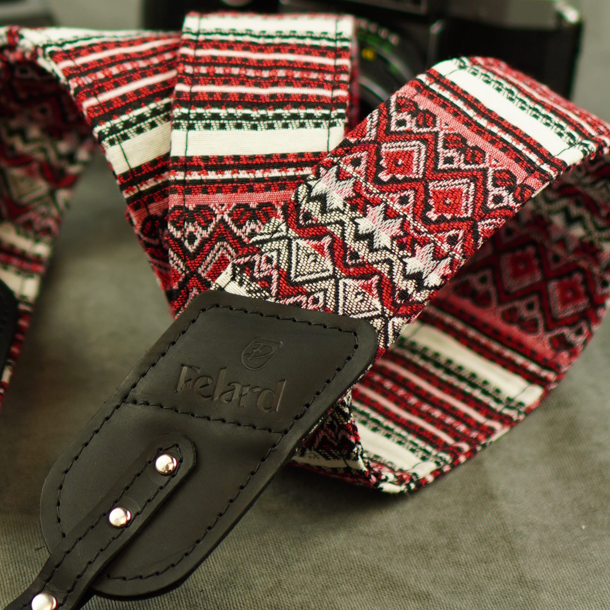 A close-up of a Felard camera strap featuring a red, white, and black geometric pattern, with a black leather brand tag.