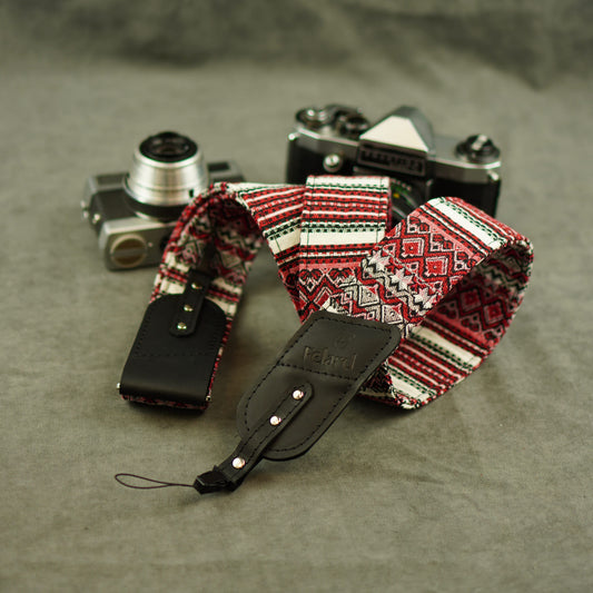 A patterned camera strap with red, white, and black geometric design and black leather ends, displayed on a gray textured surface with two vintage cameras in the background.