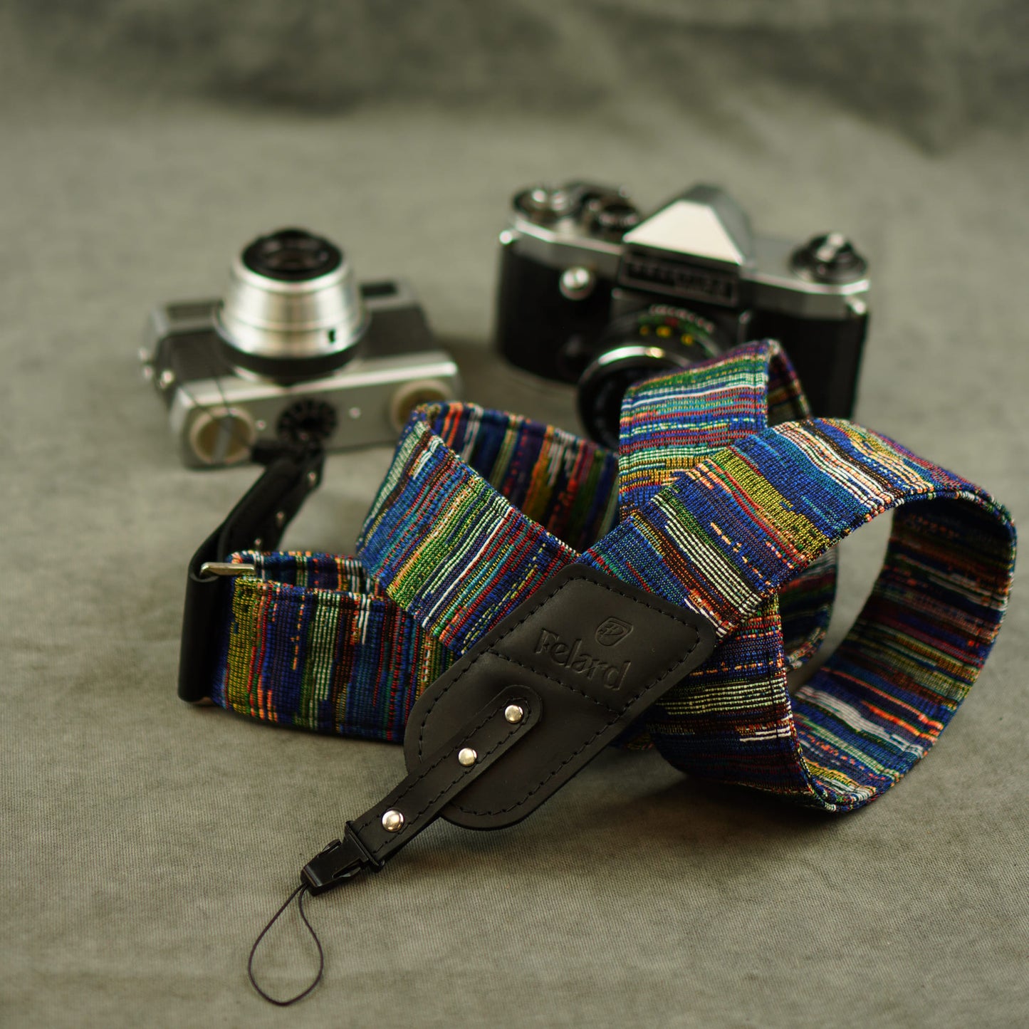 A multicolor striped fabric camera strap with a black leather displayed in front of two vintage cameras on a textured gray surface.