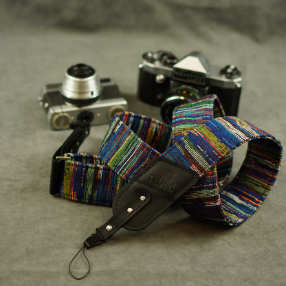 A multicolor striped fabric camera strap with a black leather displayed in front of two vintage cameras on a textured gray surface.