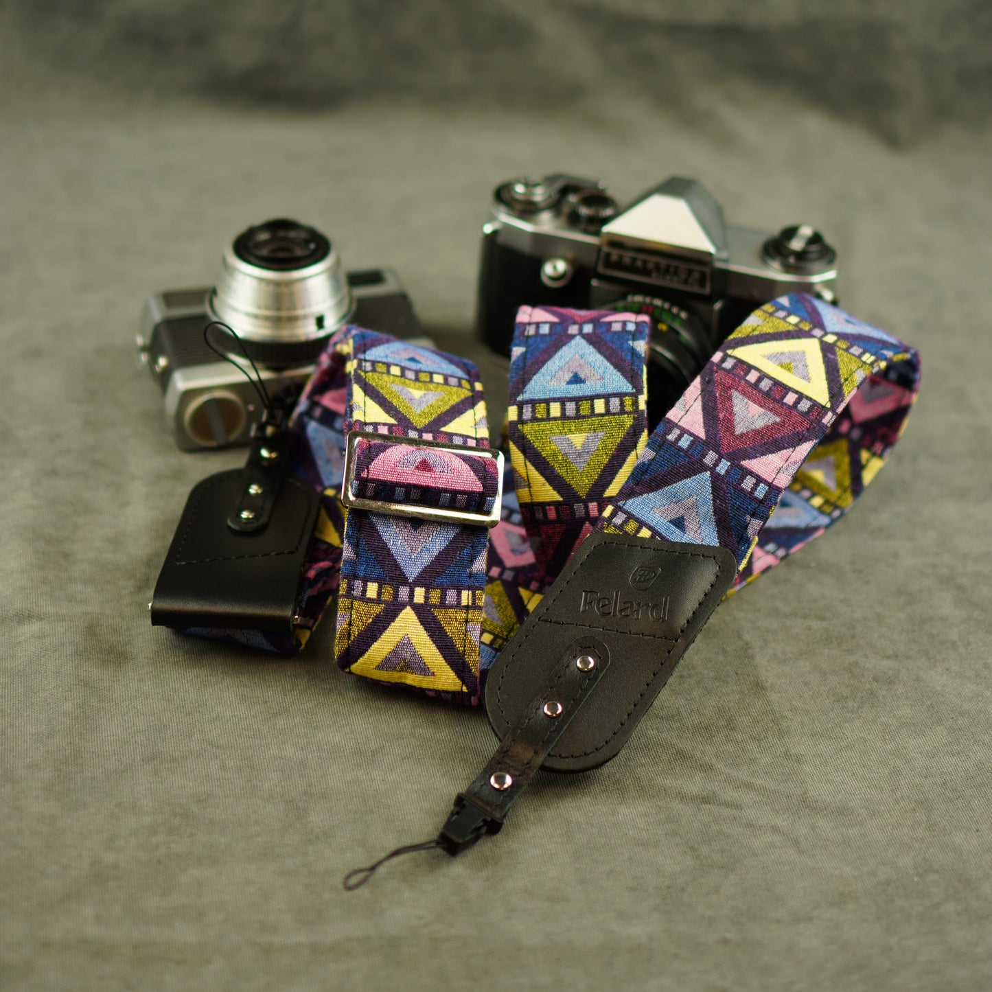 A colorful camera strap with a geometric pattern in pink, blue, yellow, and purple, shown with a black leather pouch and two vintage cameras on a textured gray background.