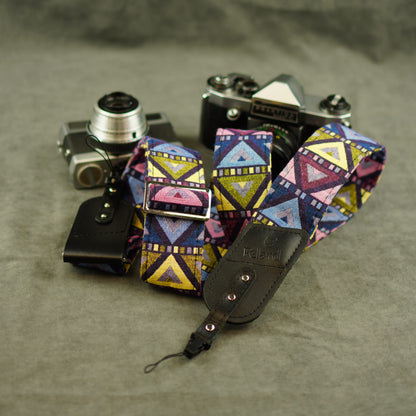A colorful camera strap with a geometric pattern in pink, blue, yellow, and purple, shown with a black leather pouch and two vintage cameras on a textured gray background.