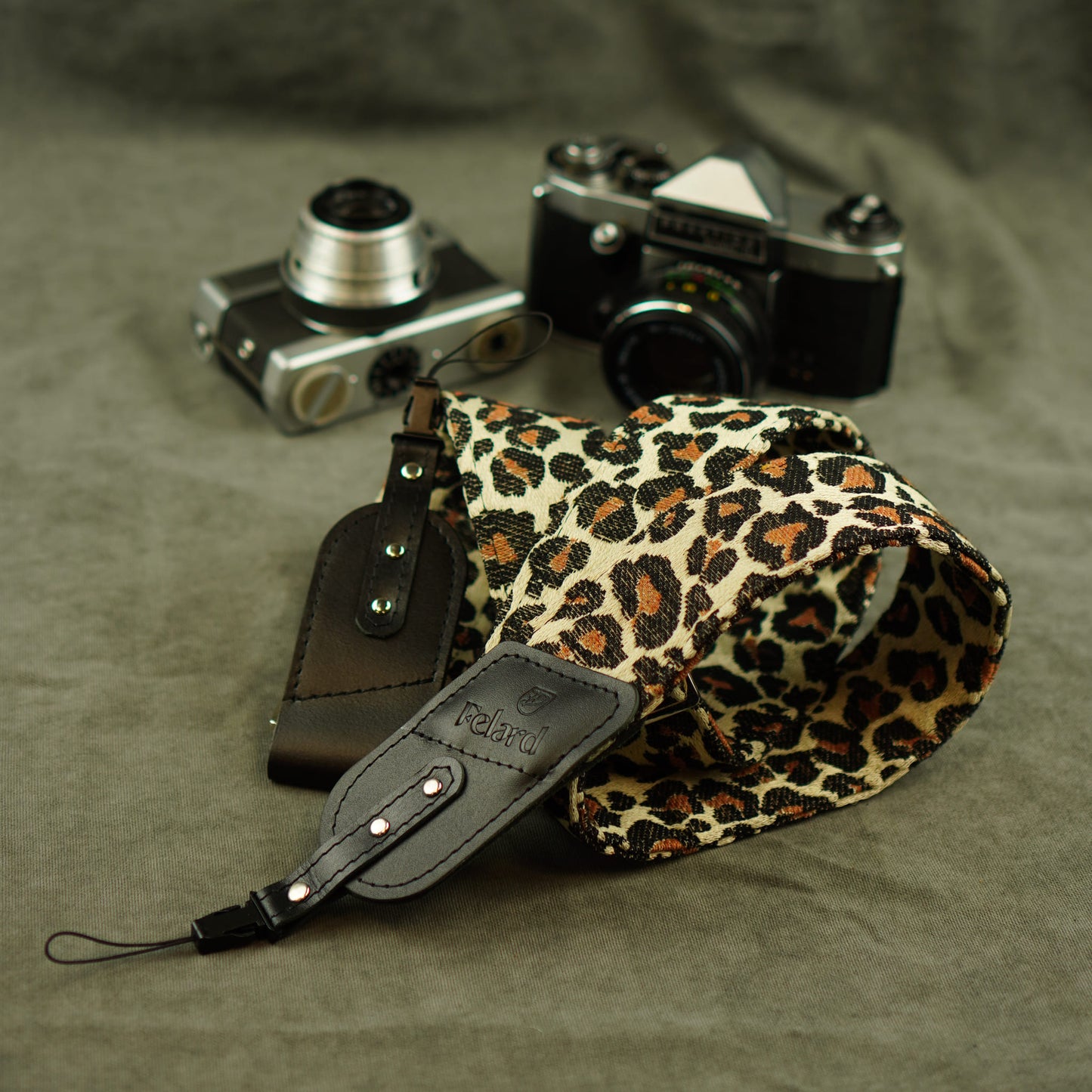 A leopard-print camera strap with black leather and tip lies on a textured gray fabric surface, styled with two vintage cameras in the background.