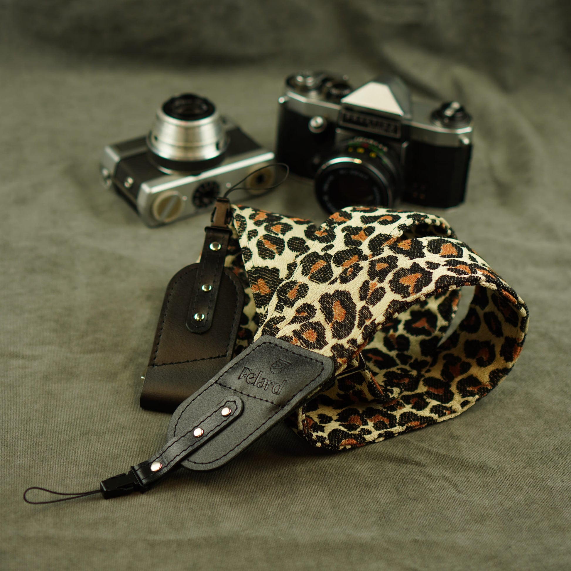 A leopard-print camera strap with black leather and tip lies on a textured gray fabric surface, styled with two vintage cameras in the background.