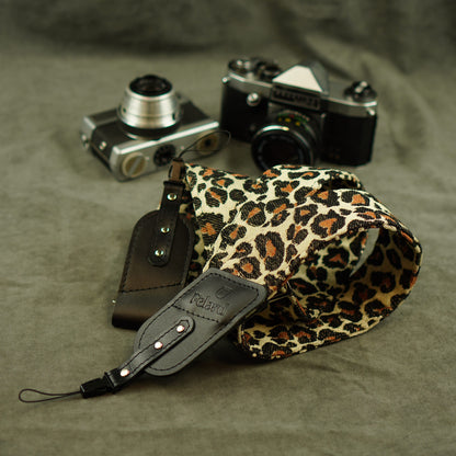 A leopard-print camera strap with black leather and tip lies on a textured gray fabric surface, styled with two vintage cameras in the background.
