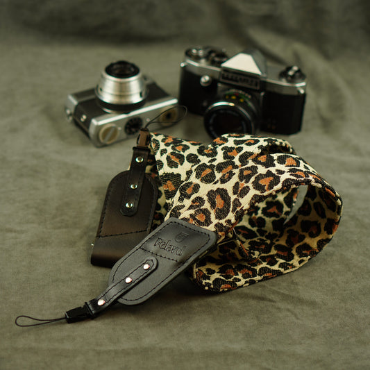 A leopard-print camera strap with black leather and tip lies on a textured gray fabric surface, styled with two vintage cameras in the background.