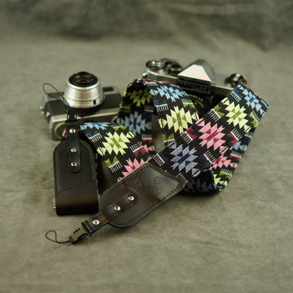 A multicolor geometric patterned camera strap with black leather attachments, shown draped over a vintage silver rangefinder camera on a textured gray surface.