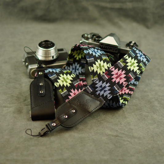 A multicolor geometric patterned camera strap with black leather attachments, shown draped over a vintage silver rangefinder camera on a textured gray surface.
