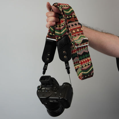 A hand holding a DSLR camera with a patterned fabric camera strap featuring a Southwestern geometric design in red, green, beige, and black, against a plain light gray background.