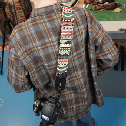 A person wearing a brown and gray plaid flannel shirt carries a camera with a colorful patterned shoulder strap. The background shows a workshop or studio setting with a workbench and tools.