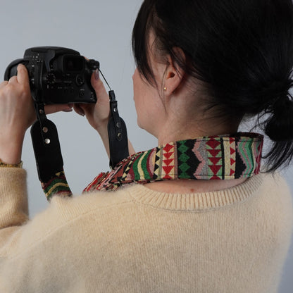 A person wearing a cream knit sweater holds a black DSLR camera with a colorful geometric patterned camera strap around their neck, shown from behind against a plain light gray background.