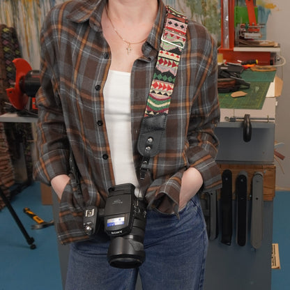 A person wearing a brown and beige plaid flannel shirt over a white tank top and blue jeans, carrying a Sony camera with a colorful geometric patterned shoulder strap in a workshop setting.
