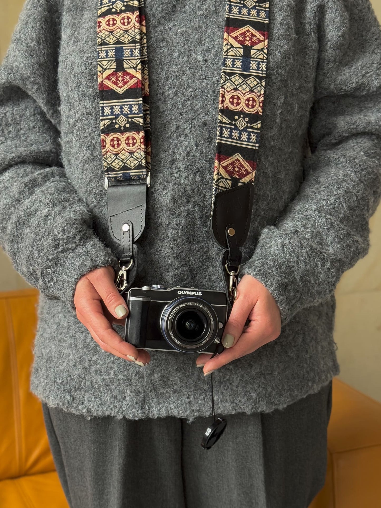 A person wearing a gray knit sweater holds a black Olympus camera with a patterned fabric camera strap featuring geometric Southwestern-style motifs in red, beige, and black.