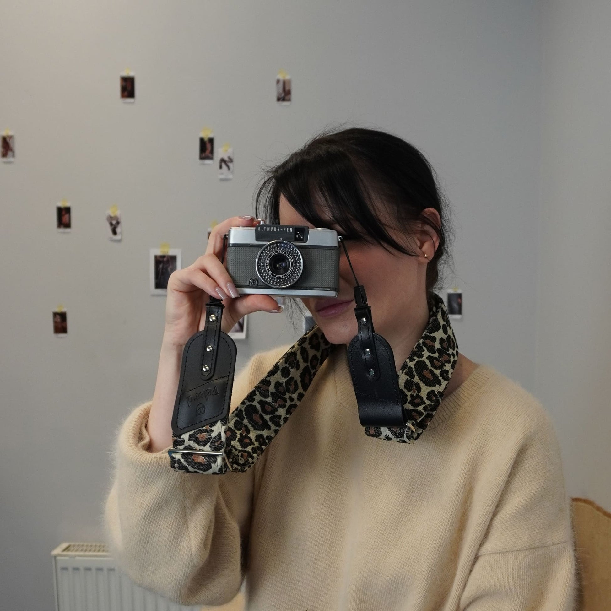 A person holds a vintage-style camera with a leopard-print and black leather neck strap in front of their face, standing against a light gray wall with small photos pinned to it.