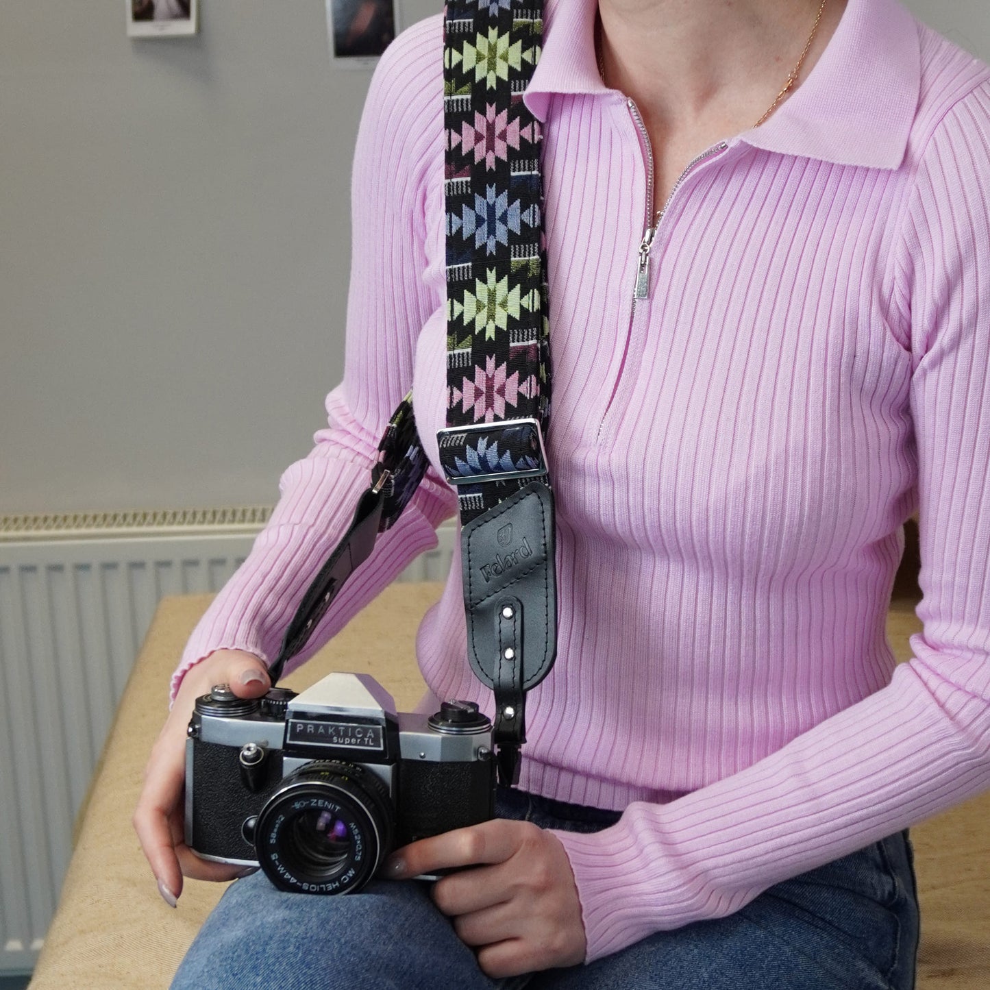 A person holds a vintage camera with a colorful geometric-patterned camera strap. The person is seated indoors next to a radiator.