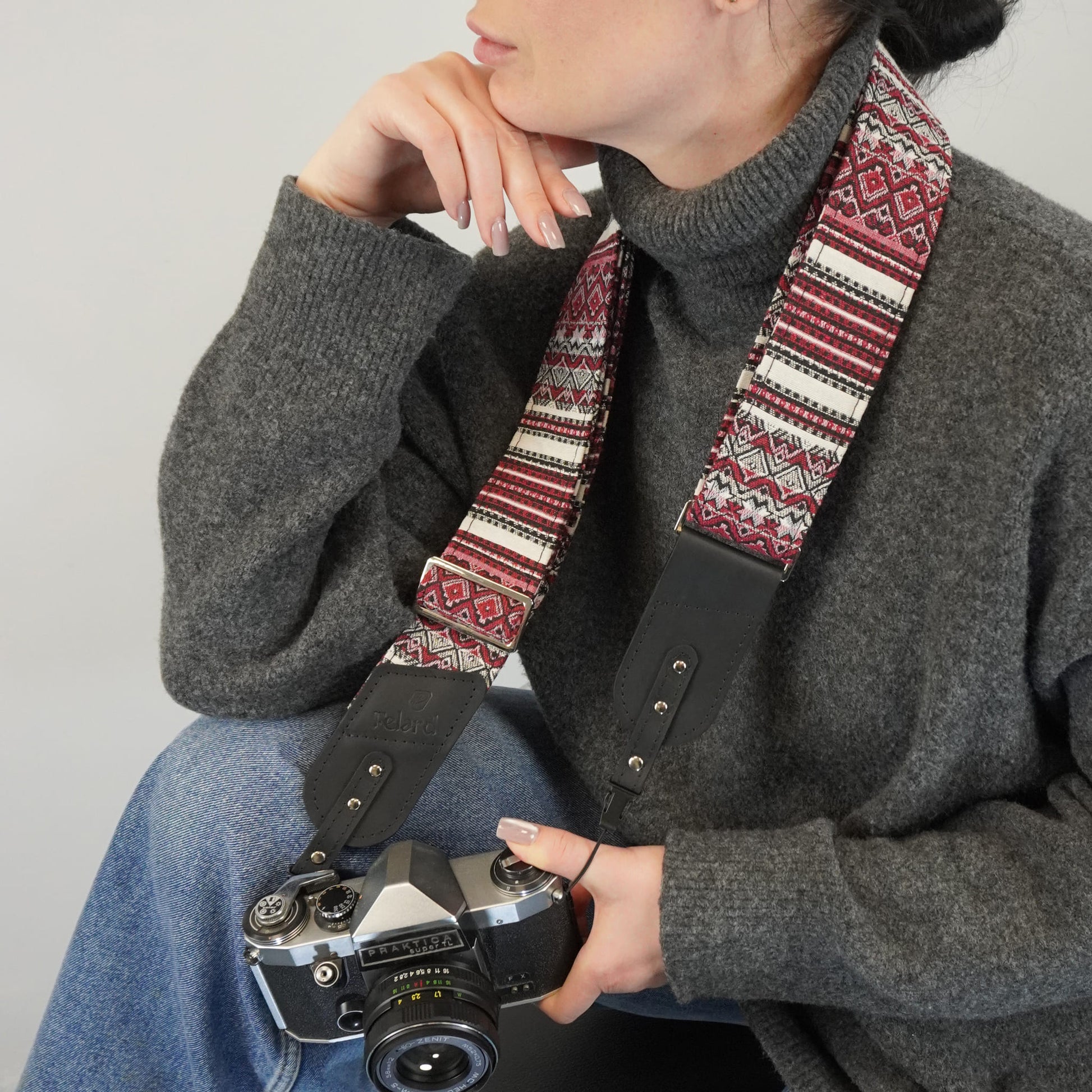 A close-up lifestyle shot of a person holding a camera with a patterned camera strap. The strap features a red, white, and black geometric Fair Isle design with black leather ends.