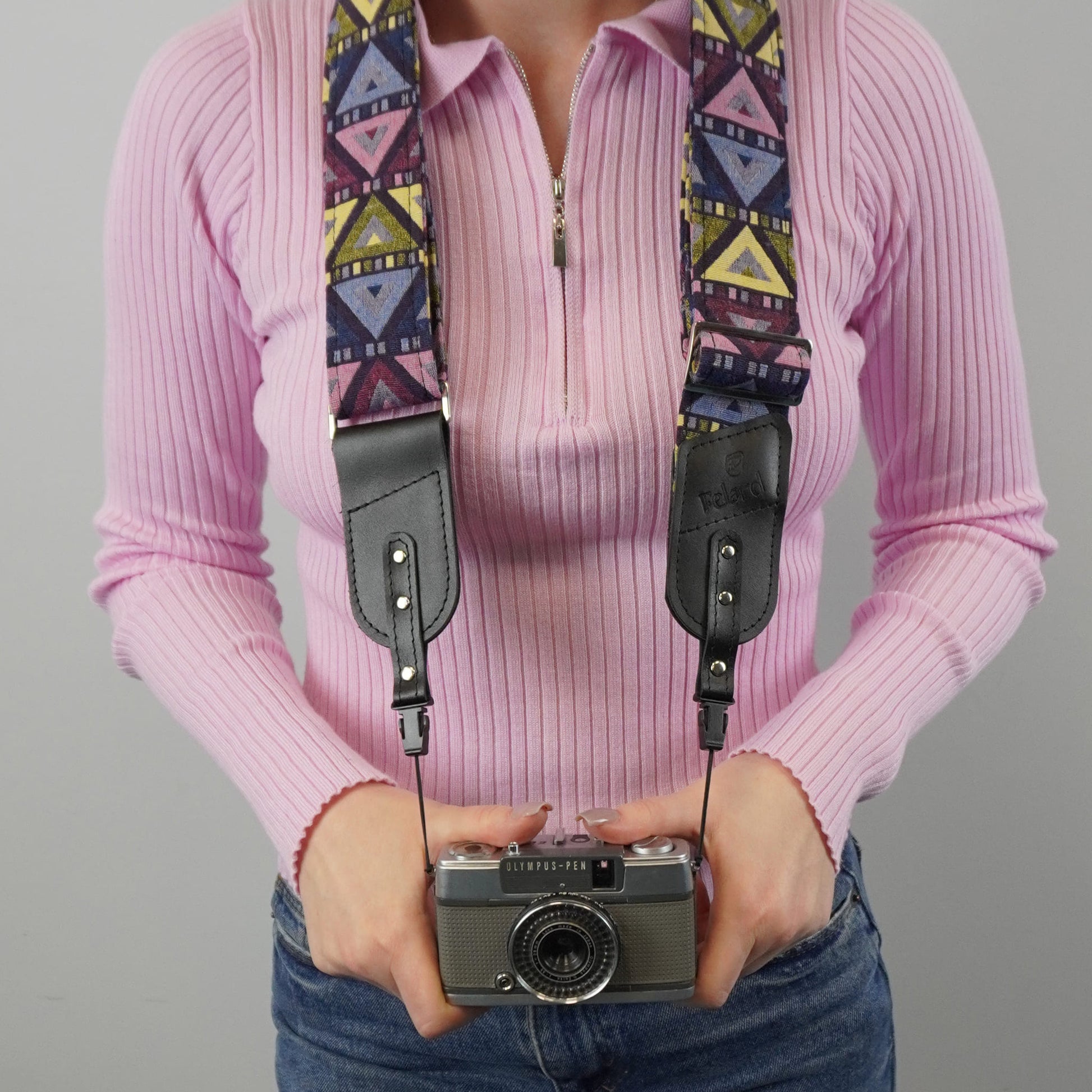 A person holds a vintage camera with a colorful geometric patterned camera strap and black leather shoulder sections against a plain light gray background.