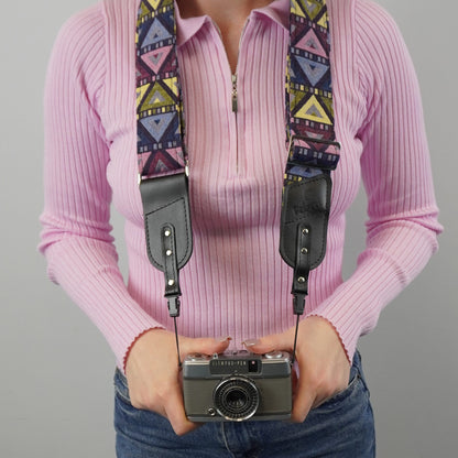 A person holds a vintage camera with a colorful geometric patterned camera strap and black leather shoulder sections against a plain light gray background.