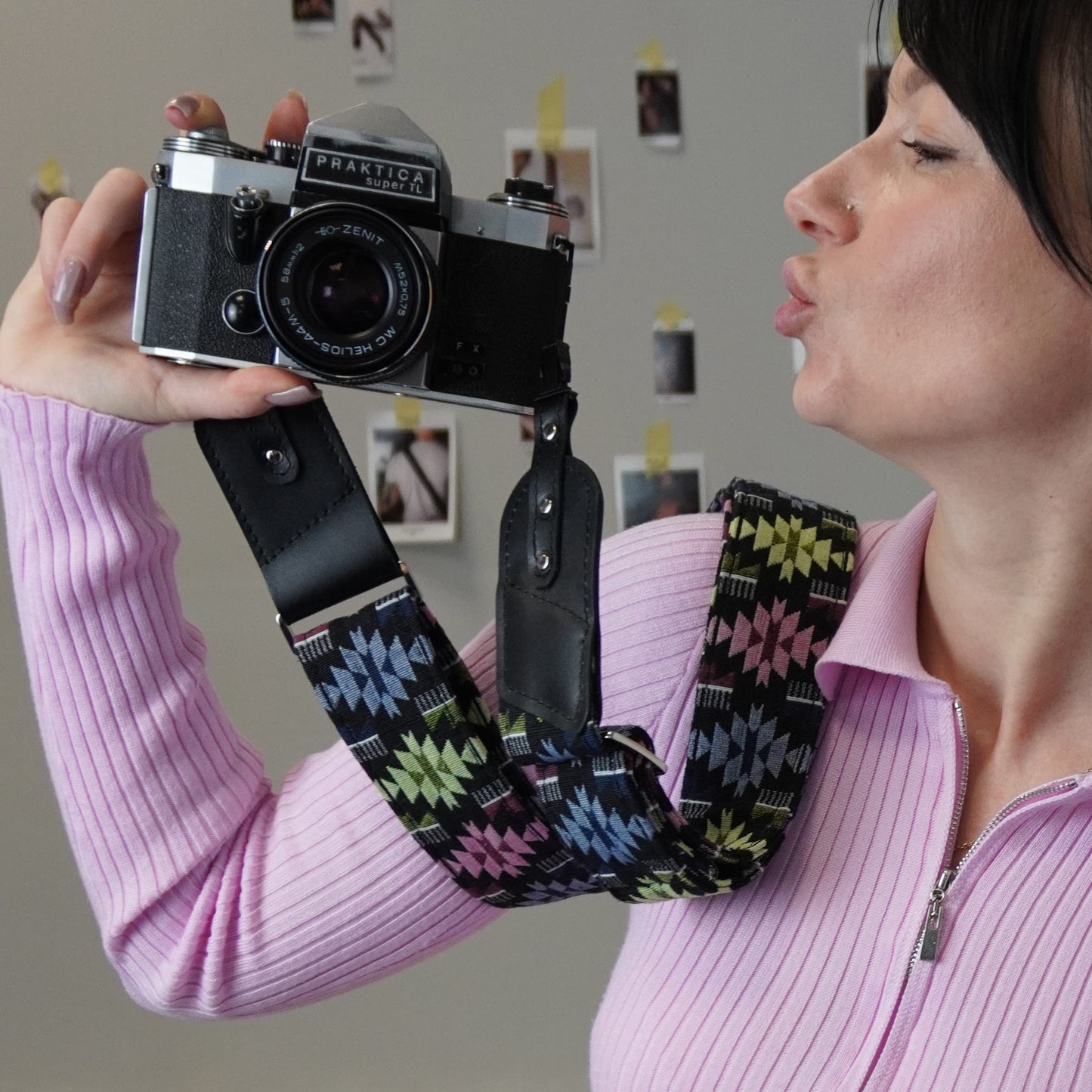 A woman holds a vintage Franca camera with a colorful geometric patterned camera strap. The strap features a black base with pink, blue, green, and yellow diamond motifs, with black leather accents.