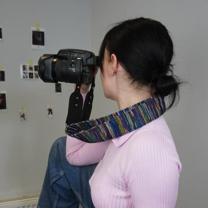 A woman in a pink ribbed top and blue jeans holds a black DSLR camera with a colorful striped camera strap around her neck, photographed from behind in an indoor setting with a light gray wall and small photos pinned on it.