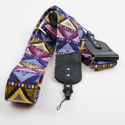 A fabric camera strap with a colorful geometric pattern in purple, pink, yellow, and blue, featuring black leather hardware and a matching pouch on a white background.