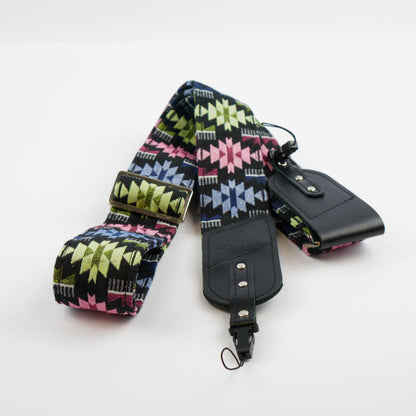 A wide fabric camera strap with a colorful geometric pattern in pink, green, blue, and black, featuring a black leather buckle and a matching black leather pouch on a white background.
