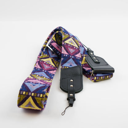 A fabric camera strap with a colorful geometric pattern in purple, blue, yellow, and pink, featuring black leather accents and a matching zippered pouch.